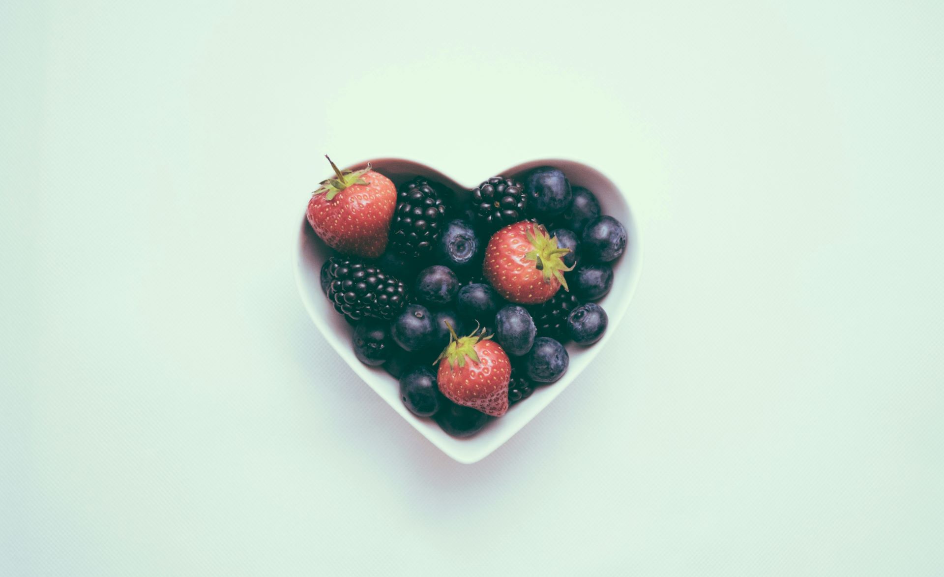 Herzförmige Schale mit frischen Beeren wie Erdbeeren, Brombeeren und Blaubeeren, dekorativ auf einem hellen Hintergrund.