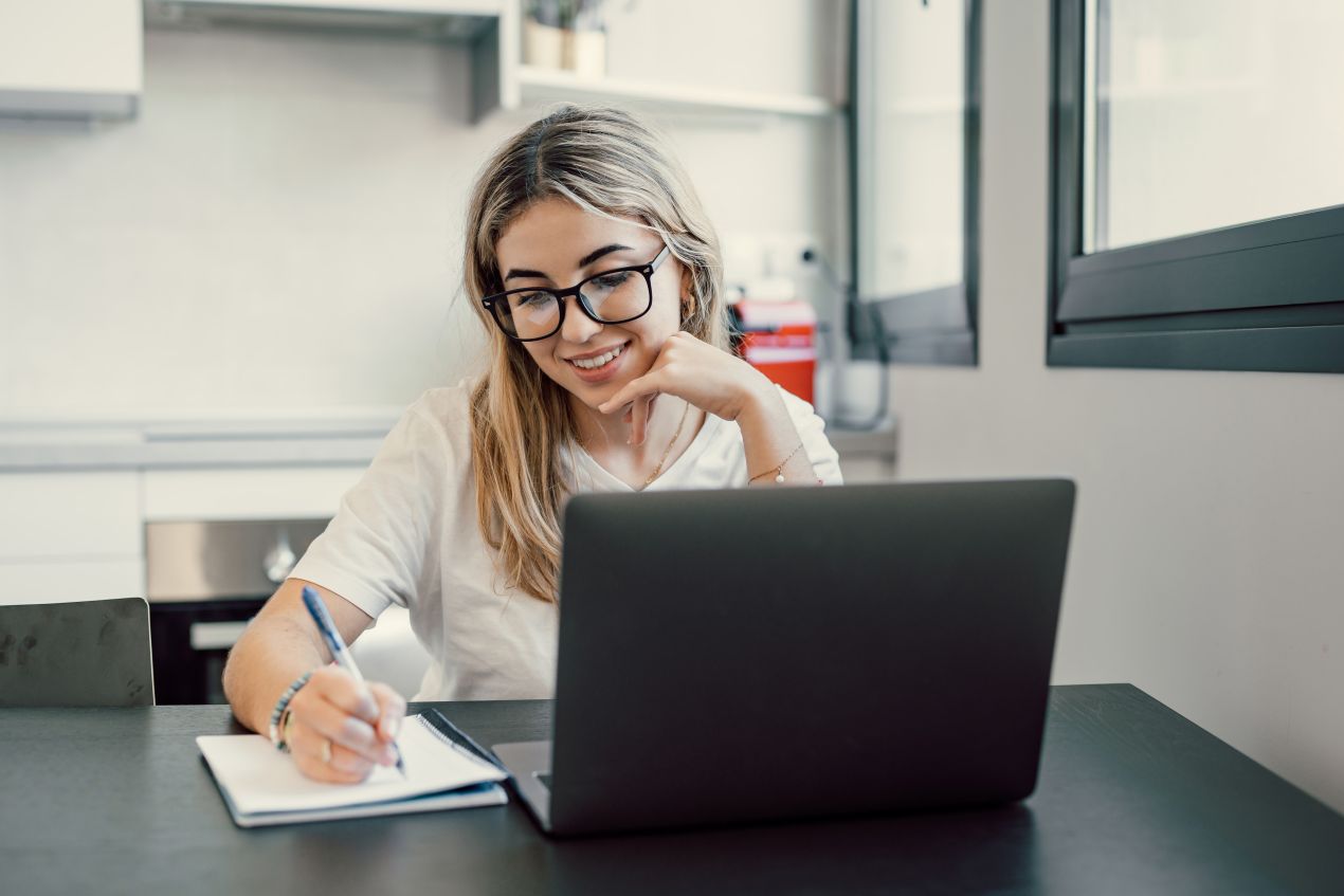 Junge Frau sitzt mit Laptop am Tisch, macht Notizen und nimmt an einem E-Learning Kurs Datenschutz teil.