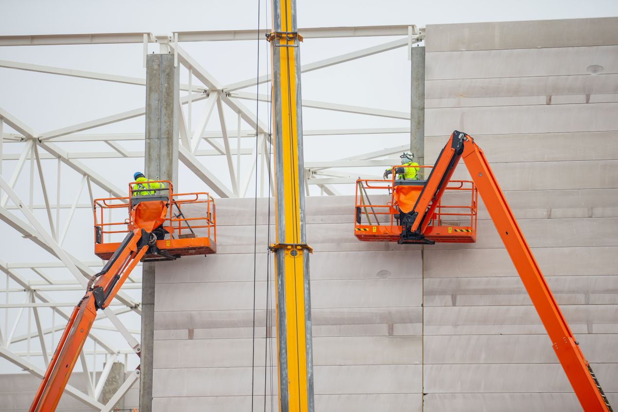 Zwei Arbeiter in orangenen Hubarbeitsbühnen montieren Bauelemente an einer Fassade, Beispiel für Schulung Hubarbeitsbühnen und sichere Arbeit auf Baustellen